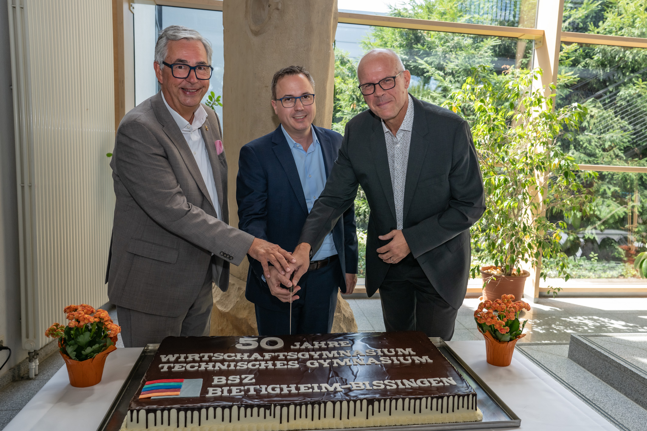 Drei Männer stehen um eine große Torte, die mit der Aufschrift "50 Jahre Wirtschaftsgymnasium Technisches Gymnasium" und einer Flagge dekoriert ist. Sie lächeln und halten gemeinsam eine Kerze über der Torte. Im Hintergrund sind Pflanzen und Fenster sichtbar.