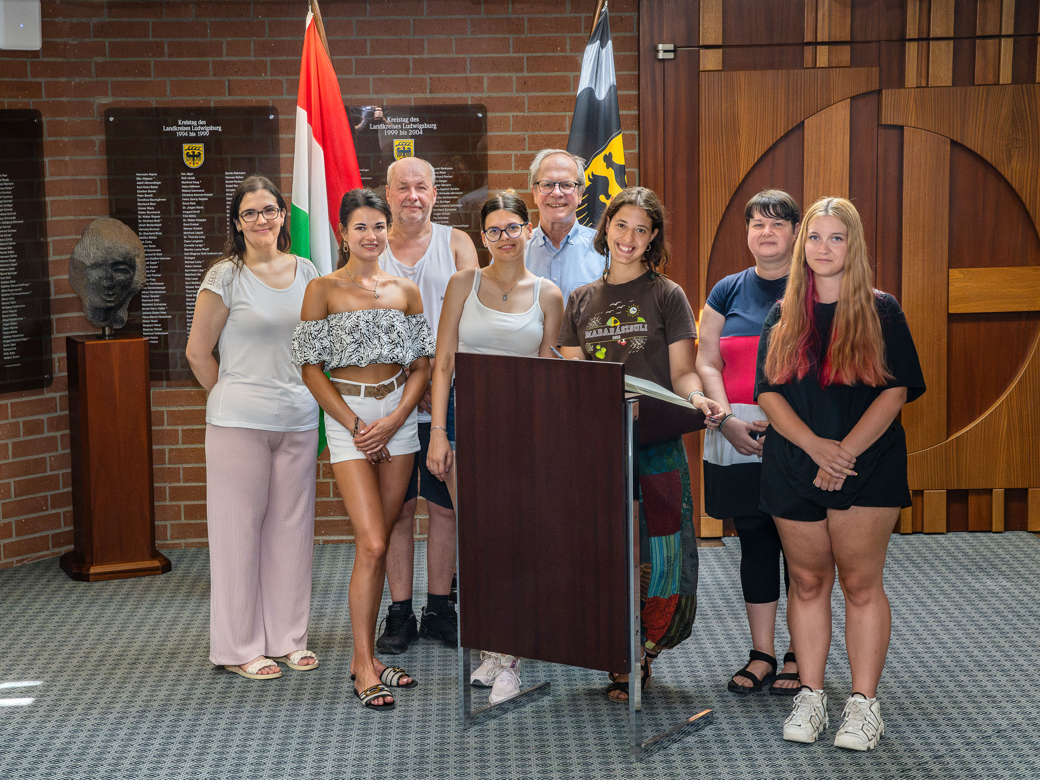 Eine Gruppe von acht Personen steht vor einem Podium in einem Raum mit Wappen und Flaggen im Hintergrund. Die Menschen lächeln und schauen in die Kamera, einige haben unterschiedliche Kleidung und Frisuren. Es scheint eine offizielle Veranstaltung zu sein. (Text KI generiert)