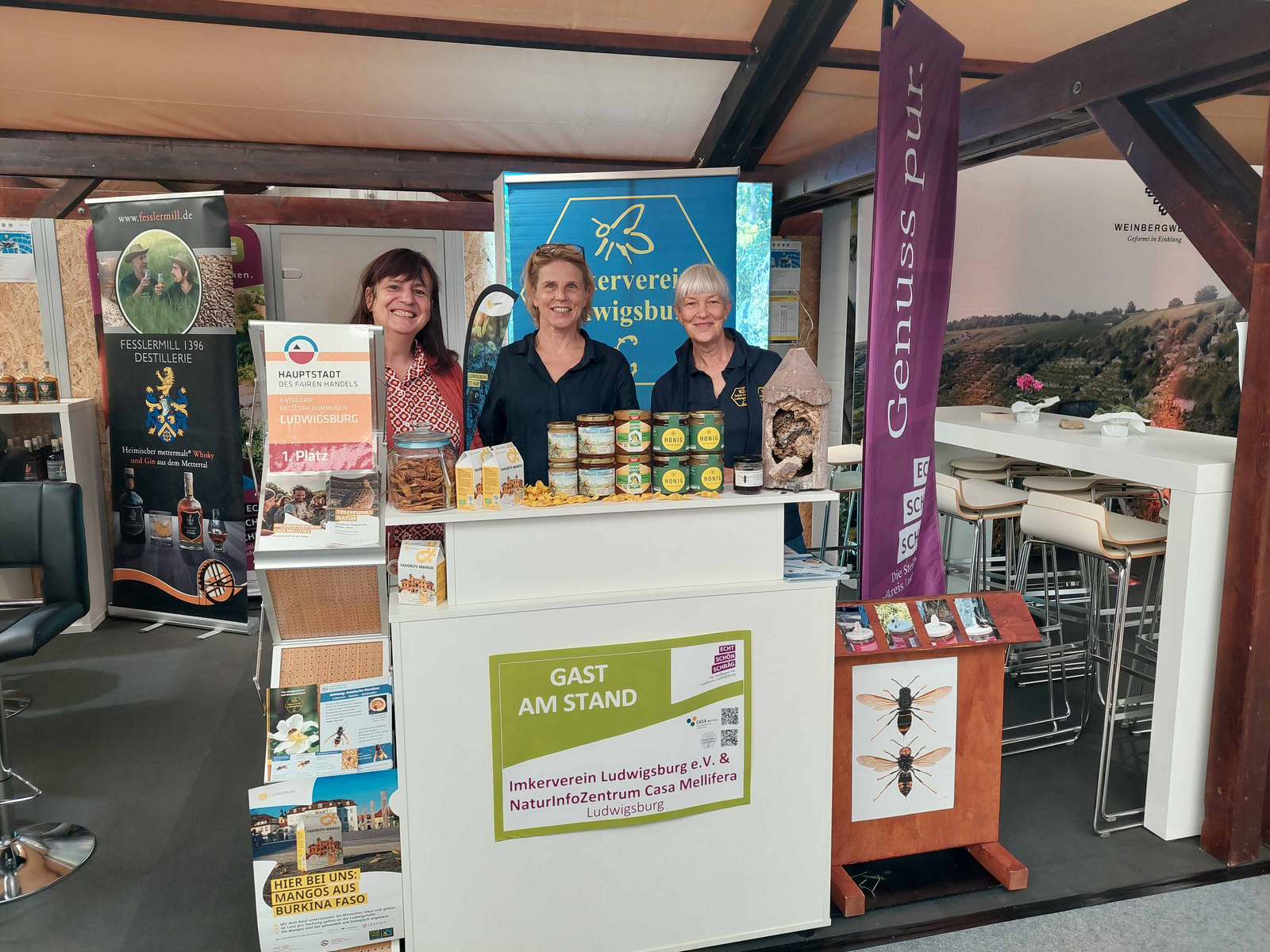Drei lächelnde Frauen stehen an einem Messestand. Sie präsentieren Naturprodukte und regionale Spezialitäten. Im Hintergrund sind verschiedene Informationsmaterialien und Banner sichtbar, die auf den Verein und das Naturerlebniszentrum hinweisen. Der Stand ist hell und einladend gestaltet. (Text KI generiert)