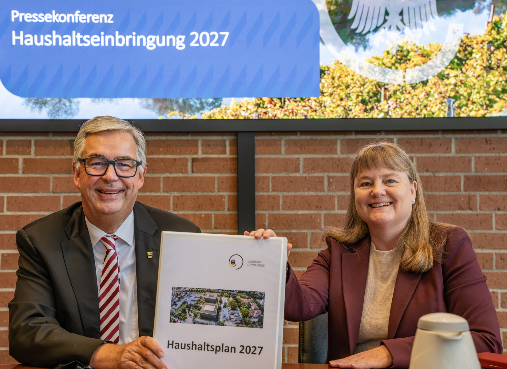 Zwei Personen sitzen an einem Tisch und präsentieren ein Dokument mit dem Titel "Haushaltsplan 2027". Im Hintergrund ist ein Banner mit der Aufschrift "Pressekonferenz Haushalts einbringung 2027" zu sehen. Beide Personen lächeln und sind formell gekleidet. (Text KI generiert)