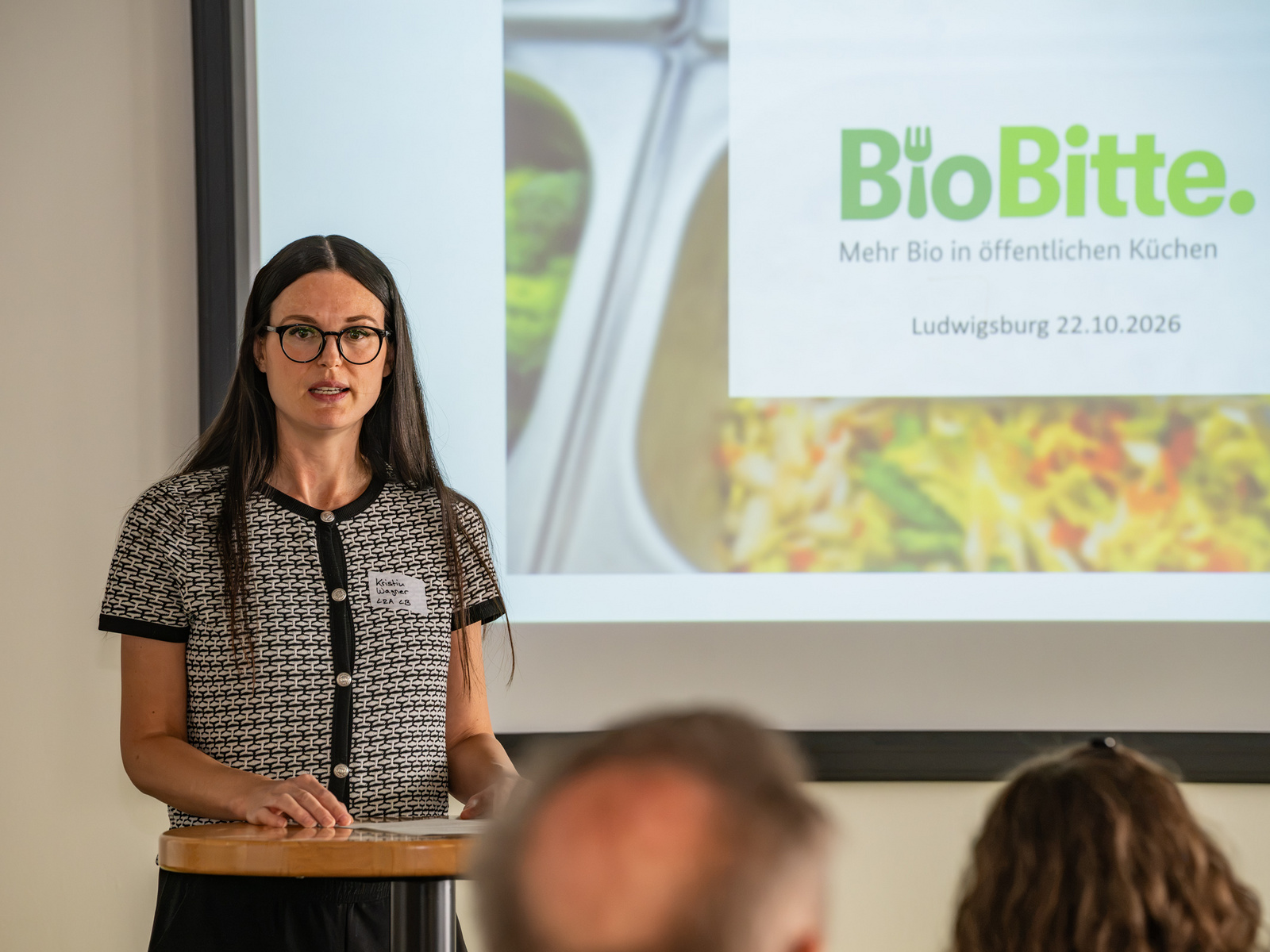 Eine Frau steht an einem Podium und spricht vor einem Bildschirm, auf dem das Logo von "BioBitte" und Informationen über eine Veranstaltung in Ludwigsburg angezeigt werden. Sie trägt eine Schwarz-Weiß-Oberbekleidung und hat lange, dunkle Haare. (Text KI generiert)