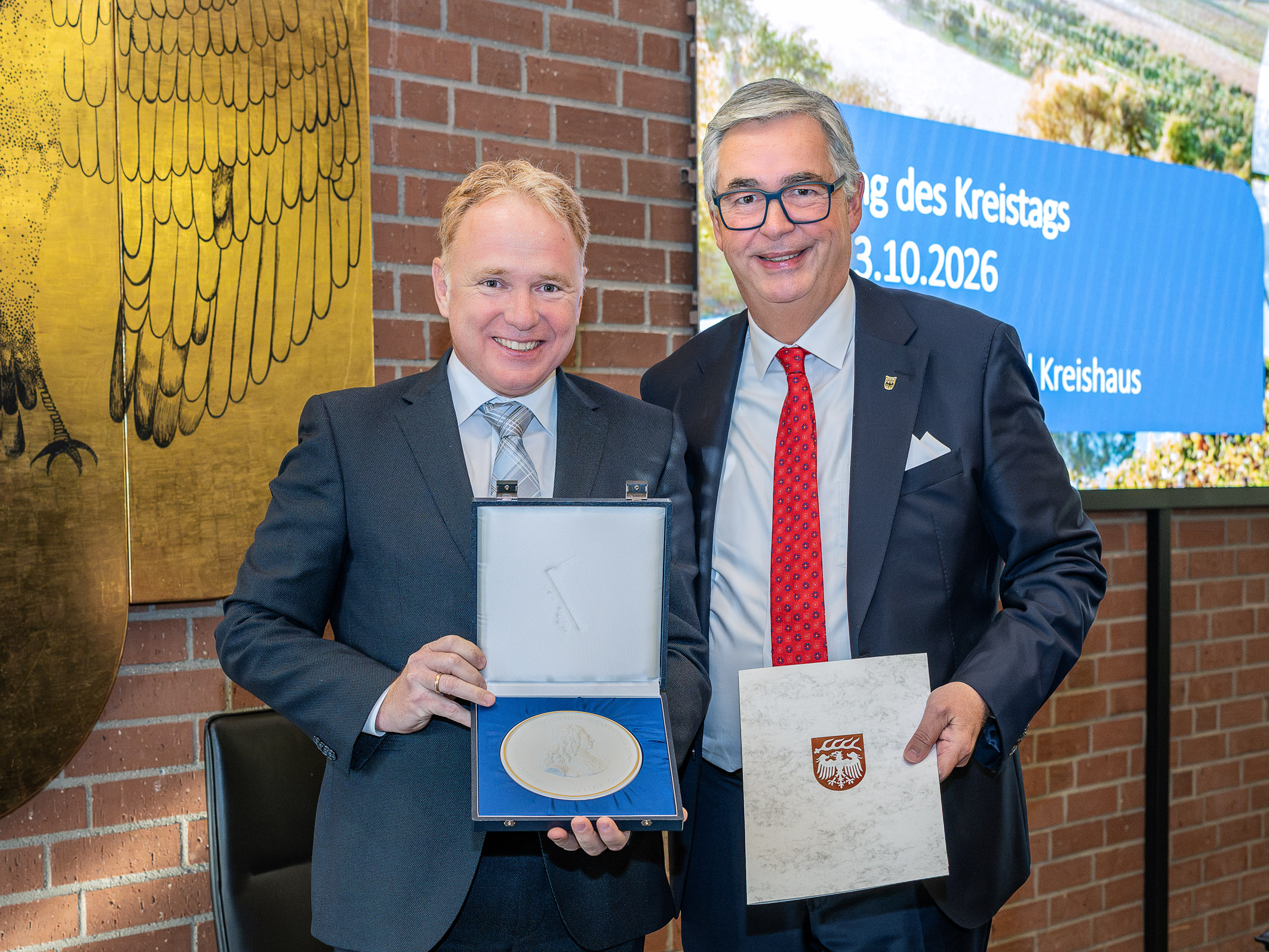 Zwei Männer stehen in einem Raum, lächeln und halten eine Medaille sowie ein Dokument. Der Mann links trägt ein hellblaues Hemd und einen dunklen Anzug, der rechts trägt einen schwarzen Anzug mit einer roten Krawatte. Im Hintergrund ist eine Wand mit einer goldenen Adlerdarstellung zu sehen. (Text KI generiert)
