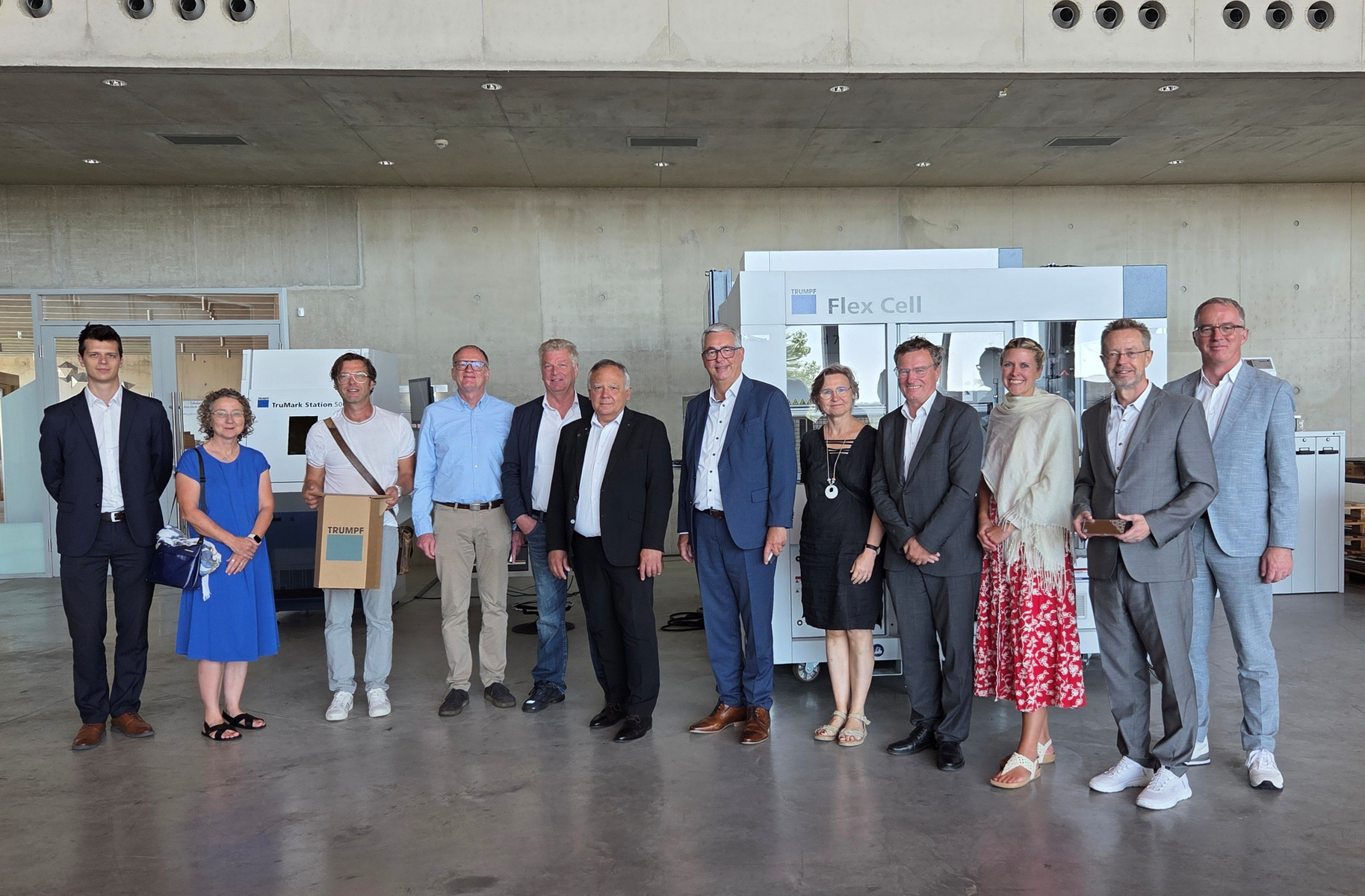 Eine Gruppe von zwölf Personen steht in einem modernen Innenraum. Sie lächeln und posieren gemeinsam für das Bild. Im Hintergrund sind verschiedene Ausstellungsstände sichtbar, darunter ein "Flex Cell"-Stand. Die Stimmung wirkt freundlich und einladend.