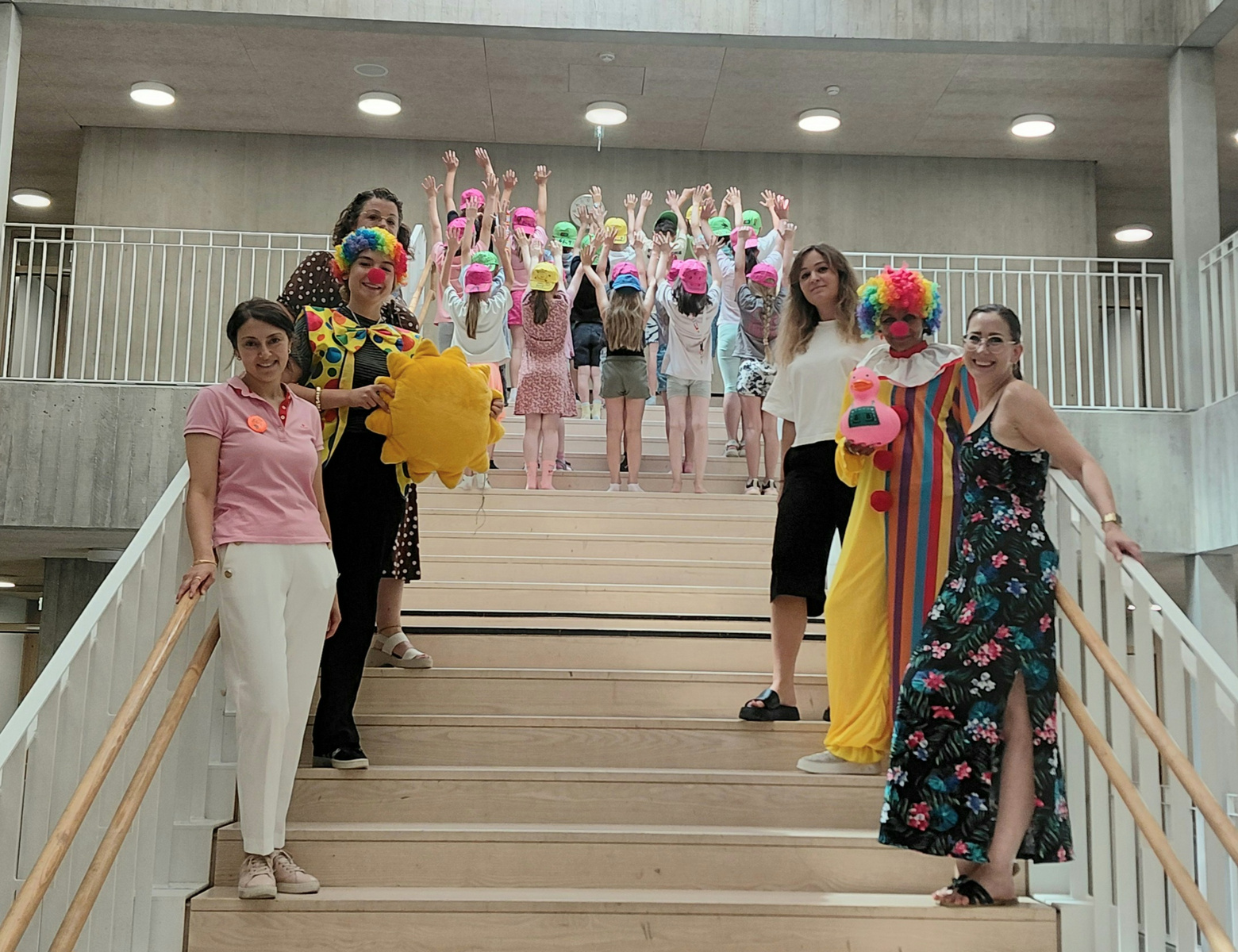 Eine Gruppe von Erwachsenen steht auf einer Treppe, während im Hintergrund Kinder mit bunten Hüten und Gesichtsbemalung fröhlich mit erhobenen Händen stehen. Die Erwachsenen tragen verschiedene Kostüme und lächeln in die Kamera, was eine festliche Atmosphäre vermittelt.