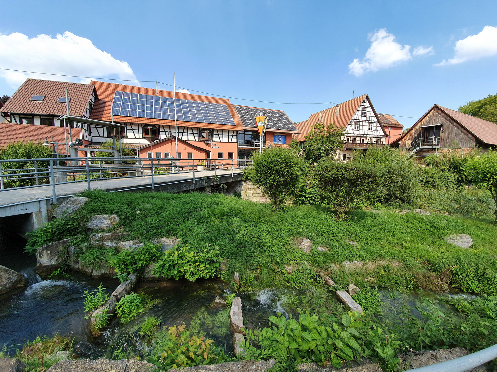 Eine malerische Ansicht eines Gebäudekomplexes mit Solarpanelen auf dem Dach, umgeben von grüner Vegetation und einem kleinen Fluss. Eine Brücke führt über den Fluss, während im Hintergrund weitere traditionelle Gebäude zu sehen sind. Der Himmel ist blau mit einigen Wolken. (Text KI generiert)
