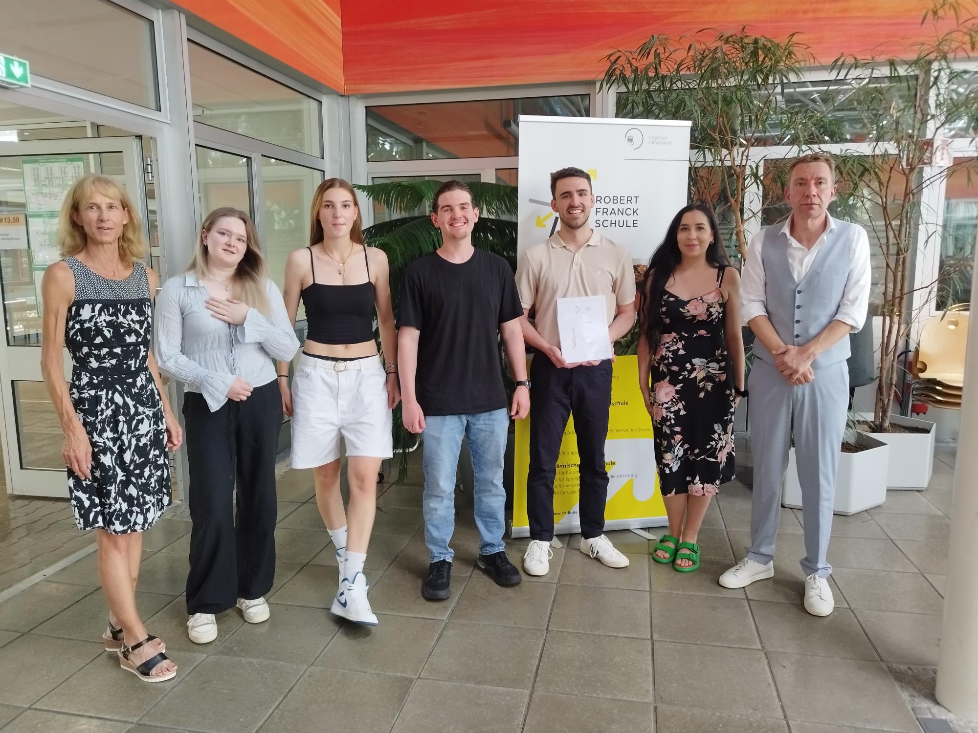 Eine Gruppe von sechs Personen steht in einem hellen Raum. Sie lächeln und posieren zusammen, während einer von ihnen ein Dokument in der Hand hält. Im Hintergrund ist ein Banner der "Robert-Franck-Schule" sichtbar. Pflanzen und große Fenster verleihen dem Raum eine freundliche Atmosphäre.