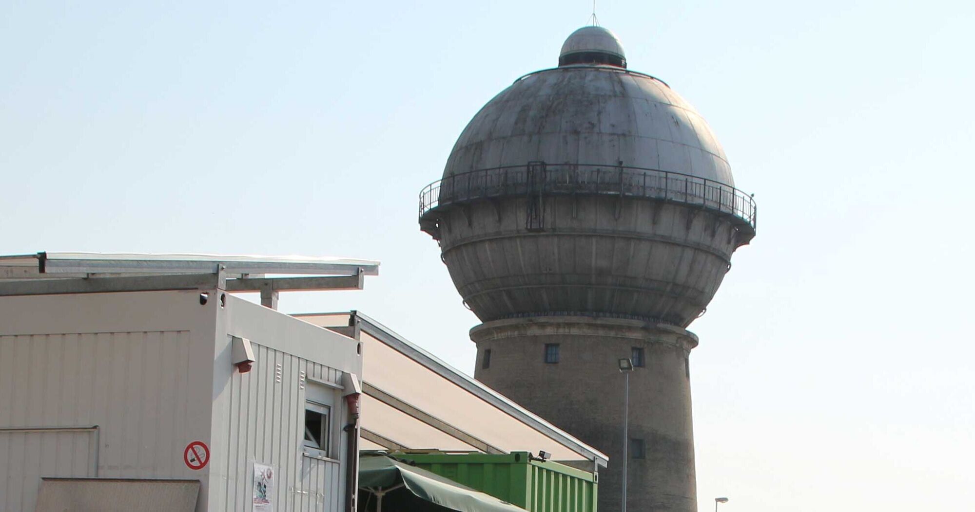Ein großer Wasserturm mit einer runden, kuppelartigen Oberseite steht neben einem niedrigeren, modernen Gebäude. Der Wasserturm hat einen grauen Farbton und eine kleine Aussichtsplattform. Der Himmel ist klar und blau im Hintergrund.