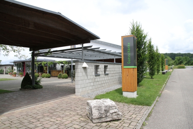 Eingang zu einem modernen Gebäude mit angrenzendem Parkplatz. Im Vordergrund eine gepflasterte Fläche und ein großes Steingewächs. Links sind gepflegte Pflanzen und rechts ein Holzschild mit der Aufschrift "Gutshof". Der Himmel ist bewölkt. (Text KI generiert)