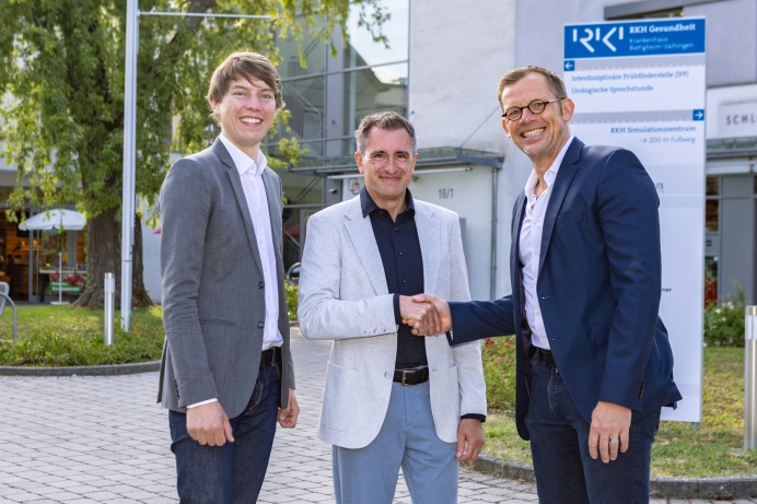 Drei Männer stehen im Freien und schütteln sich die Hände. Sie tragen formelle Kleidung. Im Hintergrund ist ein Gebäude mit einem Schild, das "RKI" anzeigt. Die Atmosphäre scheint freundlich und geschäftlich zu sein.