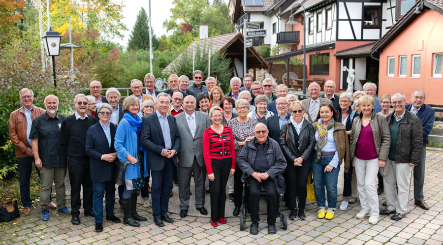 Eine Gruppe von etwa 50 Personen steht in einem Innenhof. Sie lächeln und tragen verschiedene Kleidungsstile. Im Hintergrund sind Häuser und Bäume zu sehen, die herbstliche Farben zeigen. Die Stimmung wirkt freundlich und einladend.