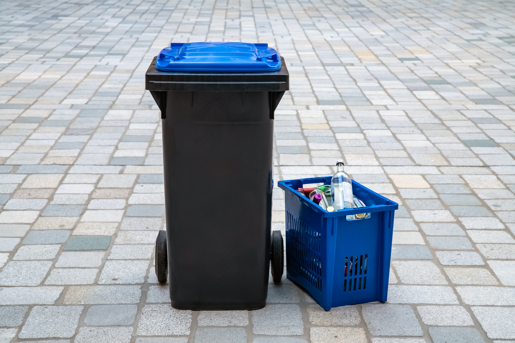 Ein schwarzer Müllcontainer mit blauer Klappe steht neben einem blauen Sammelkorb, der mit Flaschen und anderen recycelbaren Materialien gefüllt ist. Der Hintergrund besteht aus grauen Pflastersteinen.