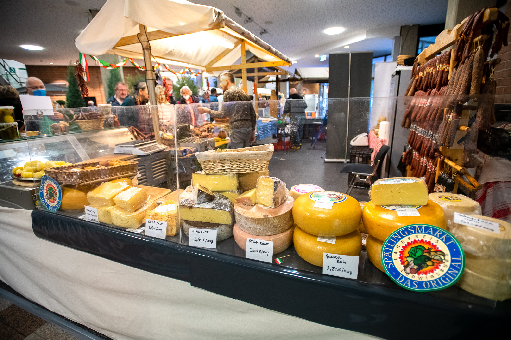 Ein Marktstand mit einer Auswahl an Käse. Verschiedene Käsesorten, die in Kästchen und Laiben präsentiert werden, sind gut sichtbar. Preise und Bezeichnungen sind an den Käsepackungen angebracht. Im Hintergrund sind Menschen zu sehen, die den Markt besuchen.