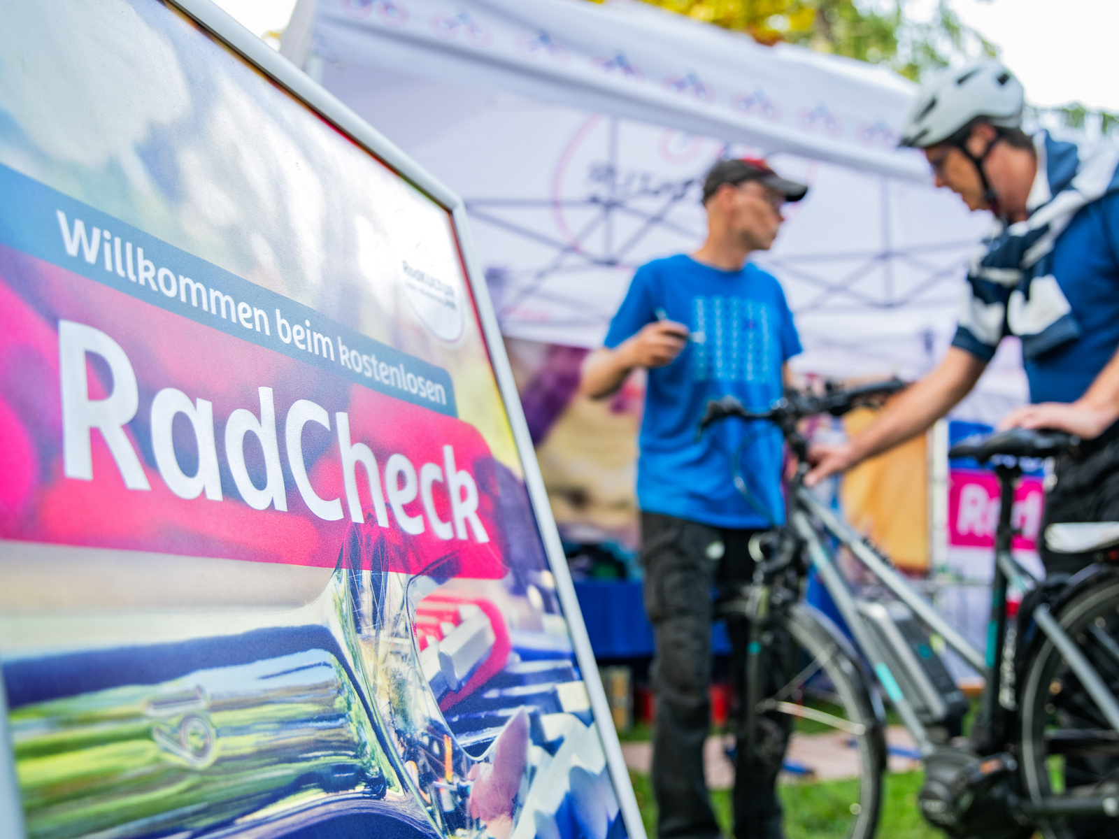 Ein Schild mit der Aufschrift „Willkommen beim kostenlosen RadCheck“ steht im Vordergrund. Im Hintergrund sind zwei Personen zu sehen, die Fahrräder überprüfen, während ein Zelt mit weiteren Informationen im Hintergrund steht.