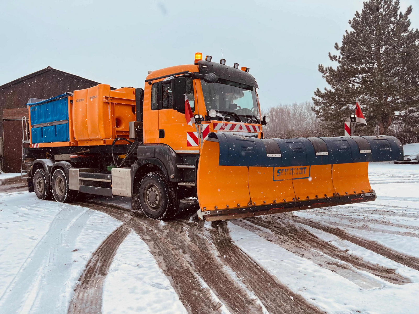 Ein orangefarbener Schneepflug steht auf einem mit Schnee bedeckten Weg. Der Lkw hat eine große, breite Schneeschaufel vorne und einen schwarzen Kasten auf der Rückseite. Im Hintergrund sind ein Gebäude und Bäume sichtbar.