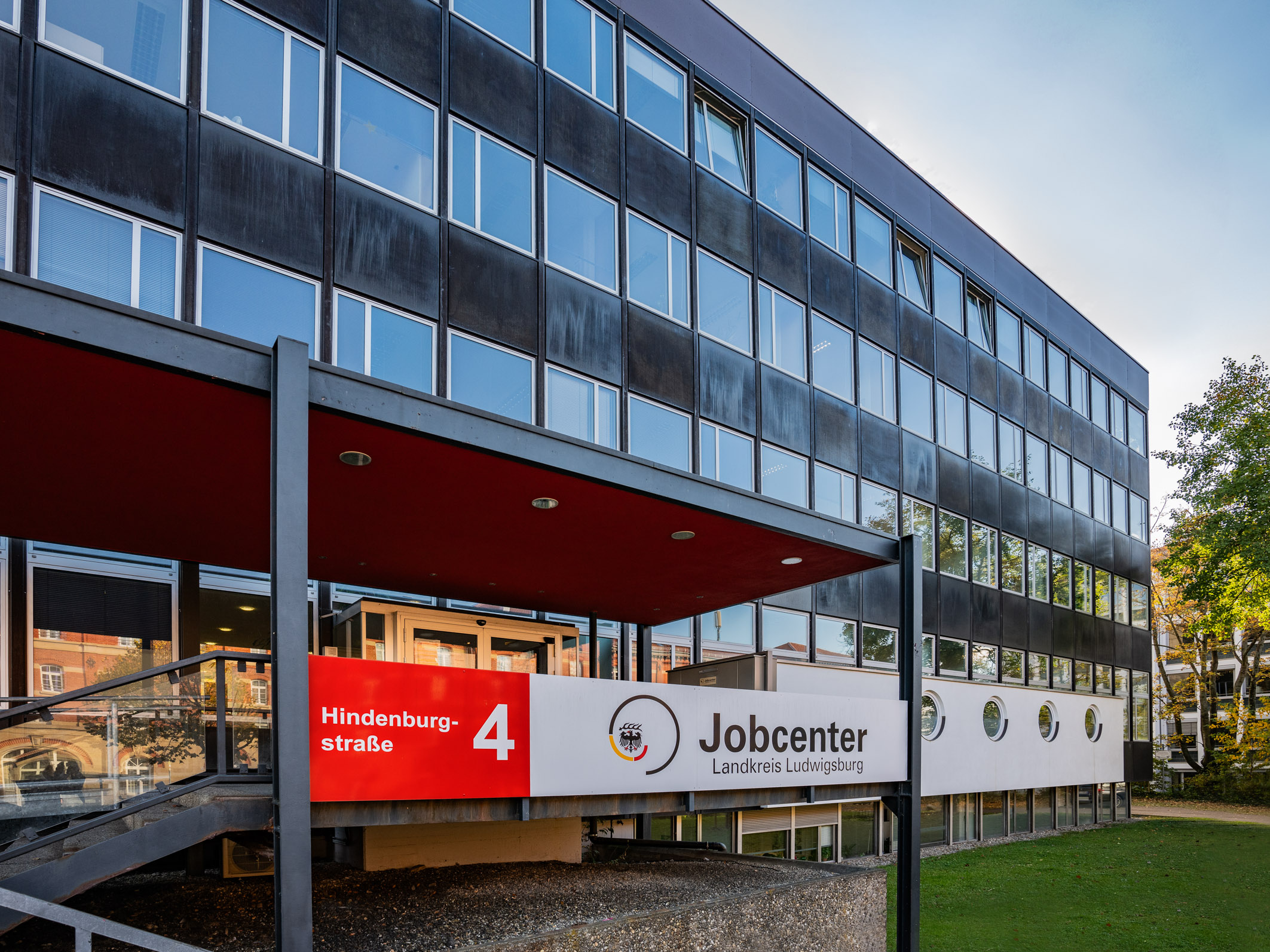 Das Bild zeigt das Gebäude des Jobcenters in der Hindenburgstraße 4. Der Eingang ist mit einer roten Überdachung versehen, und ein Schild mit der Aufschrift „Jobcenter Landkreis Ludwigslust-Parchim“ ist deutlich sichtbar. Das Gebäude hat moderne Fenster und eine gepflegte Umgebung.