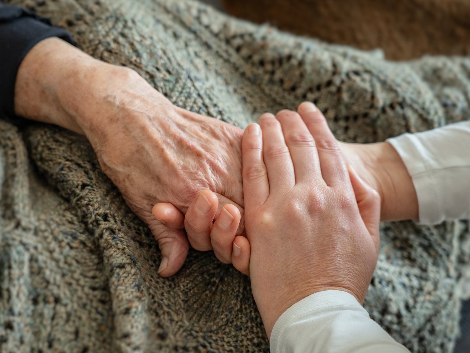 Zwei Hände halten sich liebevoll. Die eine Hand ist älter mit ausgeprägten Adern, während die andere jünger und zarter ist. Im Hintergrund ist eine Decke zu sehen. Die Geste vermittelt Wärme und Zuneigung zwischen den Generationen.