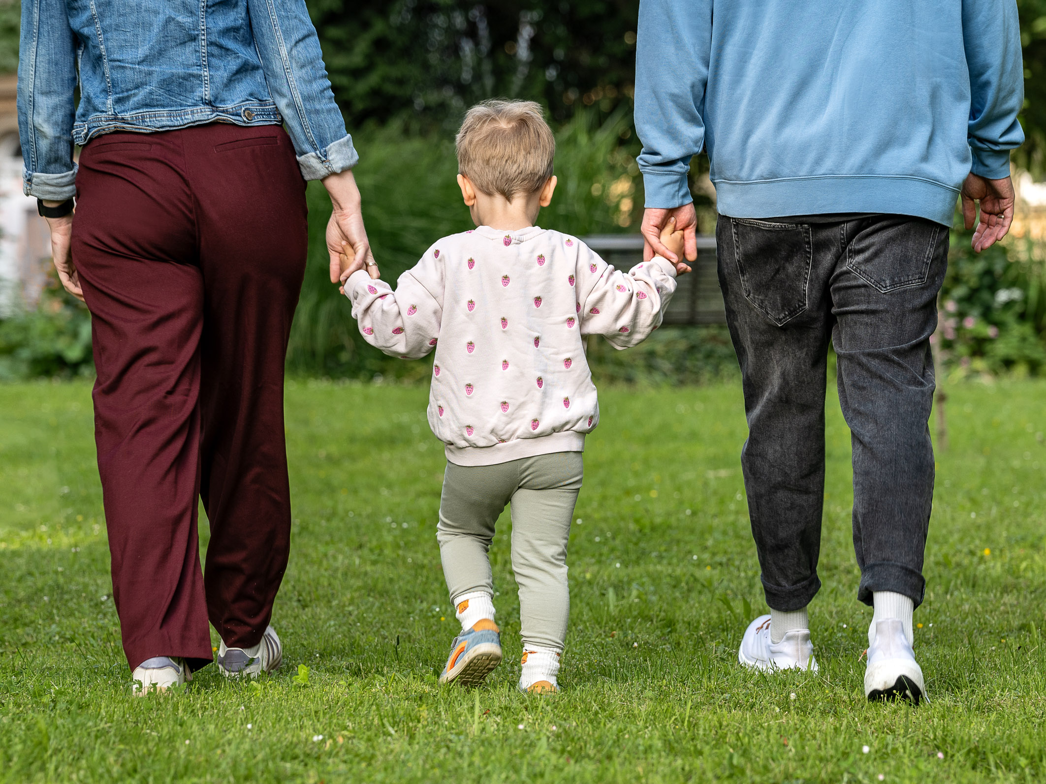 Due adulti e un bambino camminano insieme su un prato. Gli adulti tengono per mano il bambino, che indossa un maglione a pallini e pantaloni verdi. L'immagine trasmette un senso di unità e cura familiare.