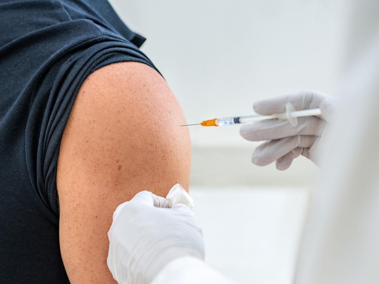 Eine Person erhält eine Impfung in den Oberarm. Eine Hand hält eine Spritze, während eine andere Hand ein Tuch bereitstellt. Der Arm ist gut sichtbar, und die Person trägt ein schwarzes T-Shirt.