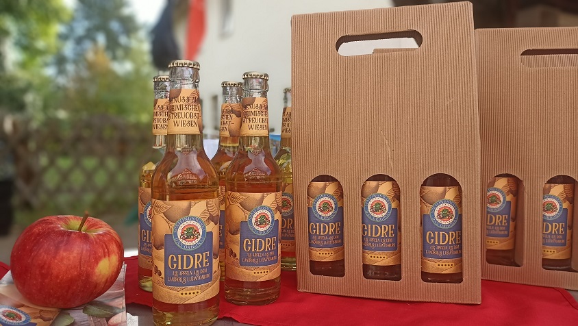 The image features several bottles of a beverage labeled "GIDRE," displayed next to a bright red apple and a cardboard six-pack carrier. The setting suggests a casual outdoor environment, highlighting the drink product.