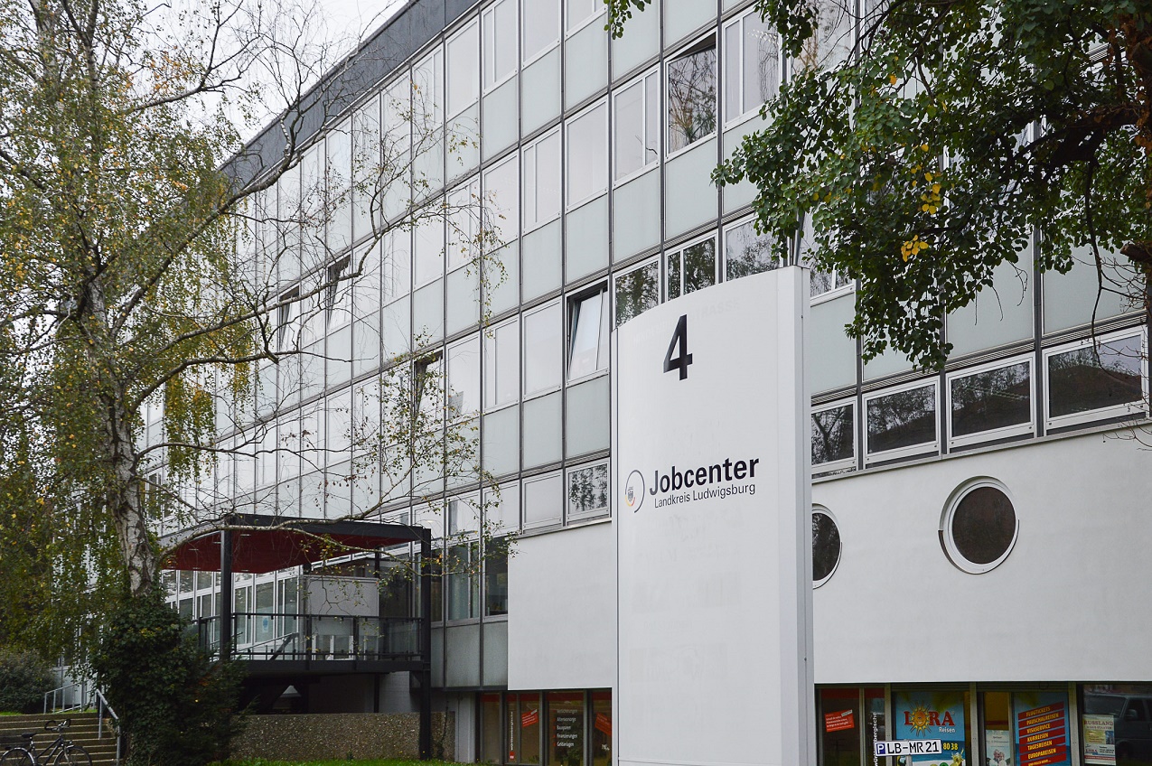 Das Bild zeigt das Gebäude eines Jobcenters in Lüneburg. Auf einem Schild ist „Jobcenter“ und die Hausnummer „4“ zu lesen. Der Eingang ist mit Pflanzen umgeben, und große Fenster prägen die Fassade des Gebäudes.