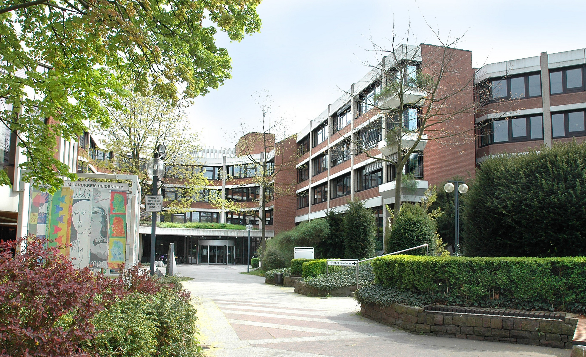 Ein weitläufiger Gebäudekomplex mit mehreren Etagen ist von Bäumen und Sträuchern umgeben. Im Vordergrund gibt es pflasterte Wege. Eine große Wandmalerei ist links zu sehen, die bunte Figuren darstellt. Der Eingang ist zentral angeordnet und einladend gestaltet.