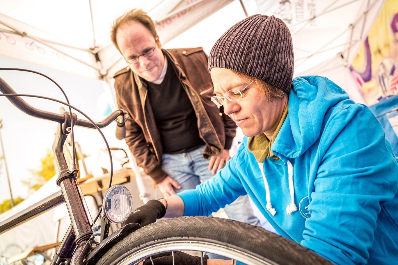 Eine Frau in blauer Jacke und mit Mütze repariert ein Fahrrad, während ein Mann im Hintergrund zuschaut. Die Frau prüft den Reifen des Fahrrads mit einem Werkzeug. Die Szene spielt sich unter einem Zelt ab, das für eine Veranstaltung eingerichtet ist.