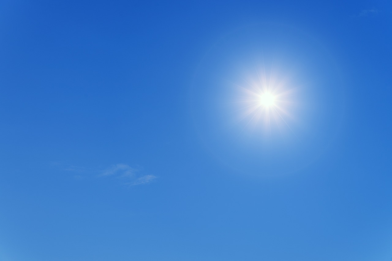 Ein klarer, blauer Himmel mit einer strahlenden Sonne. Die Sonne erscheint hell und bringt ein Gefühl von Wärme und Taglicht. Es gibt nur wenige Wolken im Hintergrund.