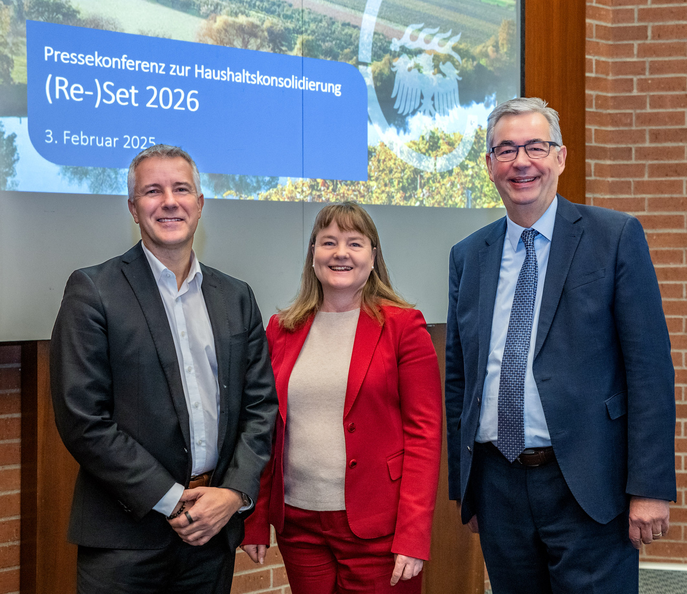 Das Bild zeigt drei Personen, die vor einer Präsentation stehen. Sie lächeln und posieren für ein Foto. Im Hintergrund ist ein Banner mit dem Text "Pressekonferenz zur Haushaltkonsolidierung (Re-)Set 2026" und dem Datum "3. Februar 2025" zu sehen.
