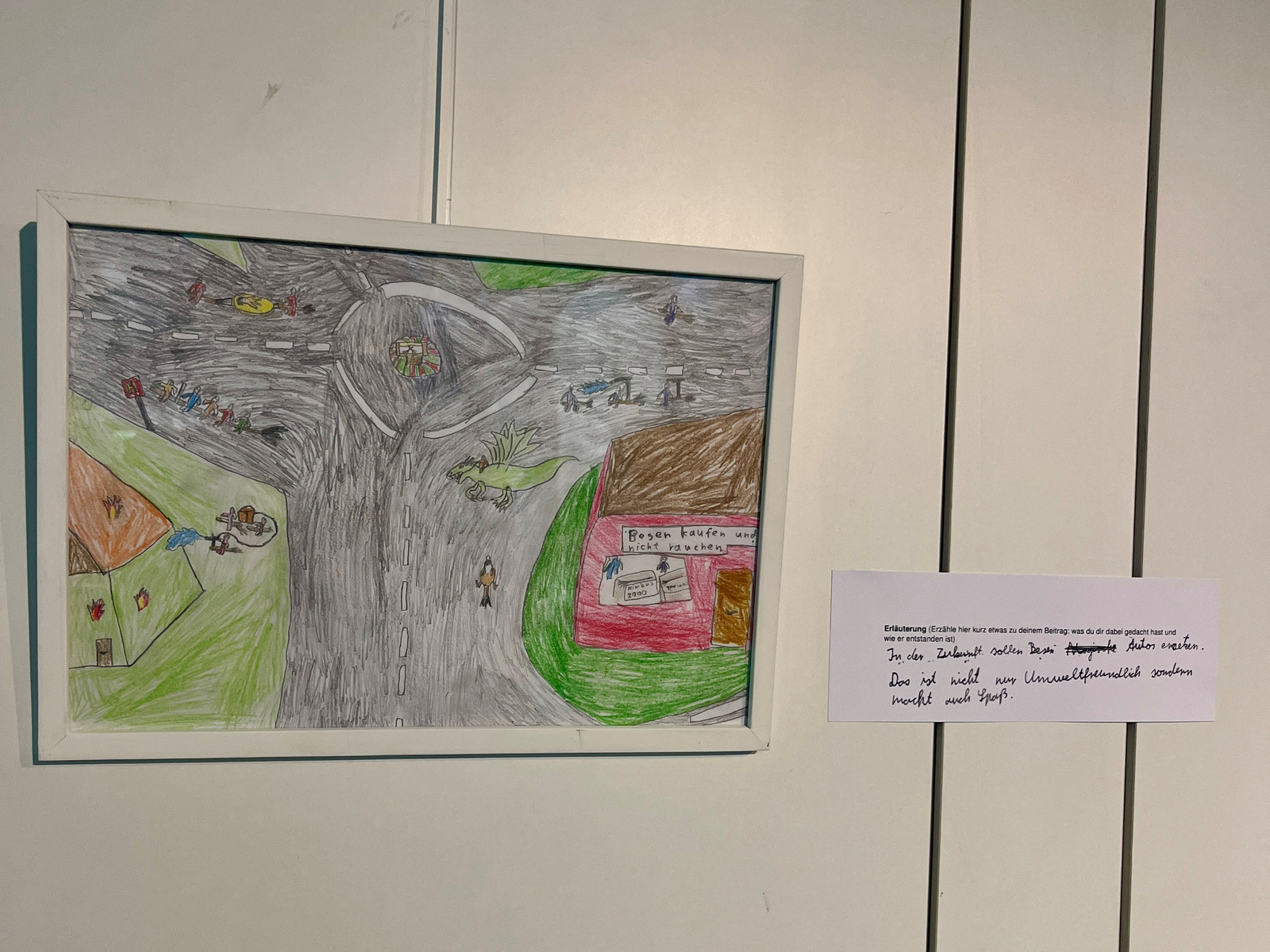 Die Zeichnung zeigt eine belebte Straßenszene mit einem Kreisel, umgeben von Häusern und Menschen, die sich bewegen. Ein Geschäft ist in der Nähe erkennbar. Die Darstellung vermittelt eine lebendige Nachbarschaftsatmosphäre. Ein erläuternder Text ist daneben angebracht.