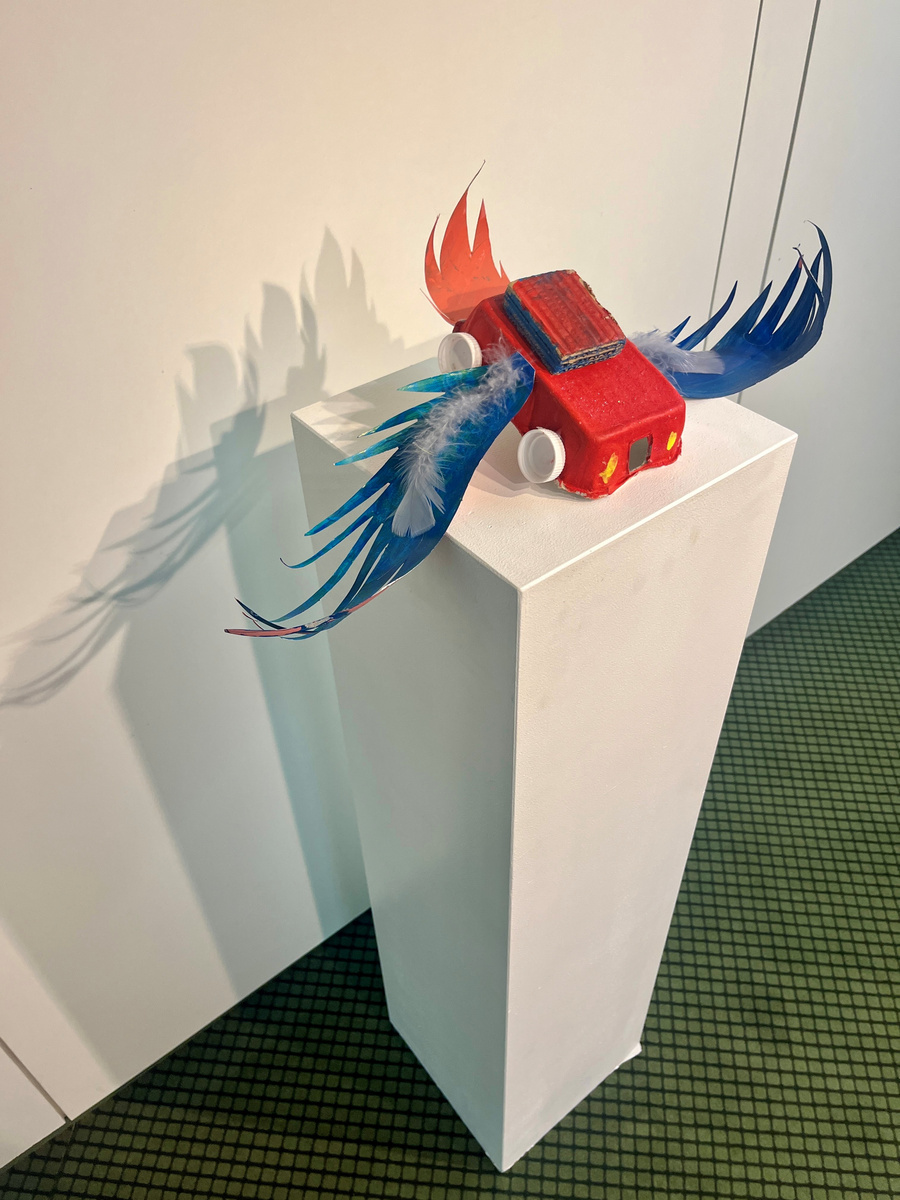 Eine rote Skulptur eines Autos mit bunten Flügeln steht auf einem weißen Podest. Die Flügel sind in Blau und Rot gestaltet und wirken lebhaft. Der Schatten des Autos und der Flügel projiziert sich auf die Wand.