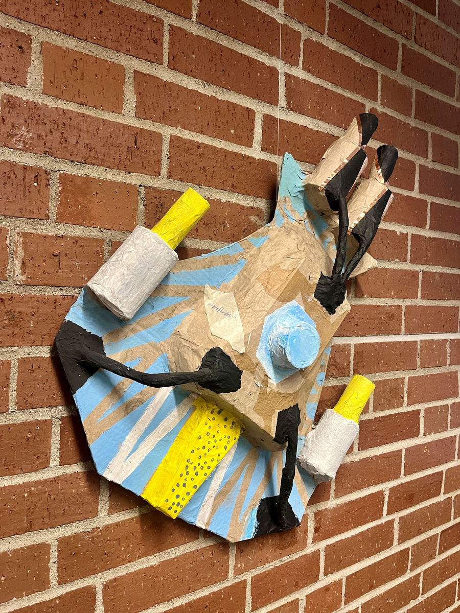 Eine künstlerische Wandinstallation in Form einer Hand, die aus Papier und farbigen Materialien gestaltet ist. Die Hand hat eine überwiegend braune Farbe, ergänzt durch blaue und gelbe Details sowie schwarze Linien, die eine lebendige, dynamische Wirkung erzeugen. Hintergrund aus roten Ziegelsteinen.