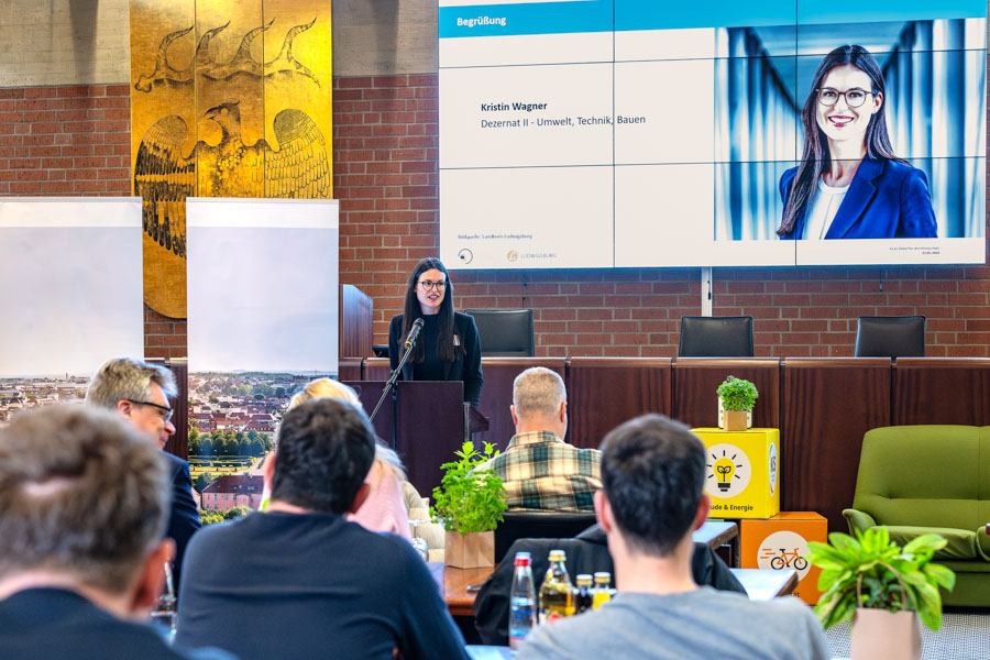Eine Frau steht am Rednerpult und spricht vor einem Publikum. Auf dem Bildschirm hinter ihr ist ihr Name und Titel zu sehen: Kristin Wagner, Dezernentin für Umwelt, Technik und Bauen. Im Vordergrund sind Teilnehmer zu erkennen, die ihr zuhören.