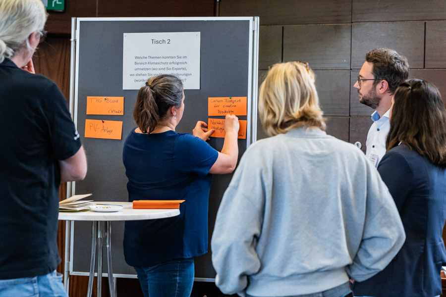 Eine Gruppe von Menschen diskutiert an einem Tisch. An einer Pinnwand hängen orangefarbene Zettel mit Themen. Eine Person klebt einen weiteren Zettel an die Wand. Oben steht "Tisch 2". Die Atmosphäre wirkt kooperativ und engagiert.