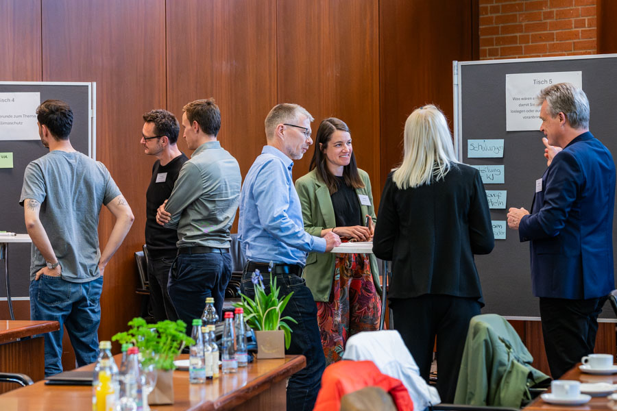Eine Gruppe von sieben Personen steht in einem Konferenzraum und unterhält sich. Einige Notizen und Plakate sind im Hintergrund sichtbar. Auf einem Tisch befinden sich Getränke und Pflanzen. Die Atmosphäre wirkt entspannt und kommunikativ.