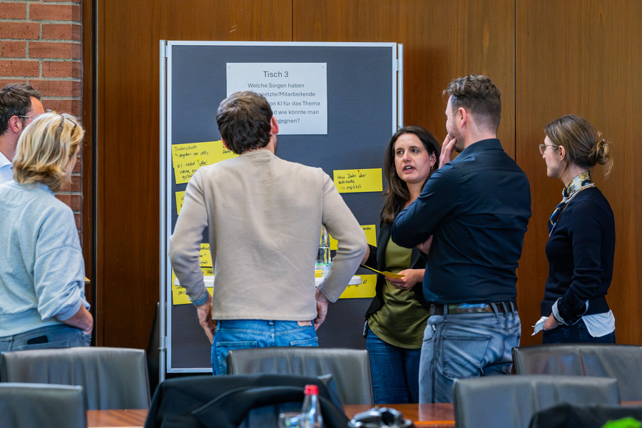 Eine Gruppe von sechs Personen steht um ein großes Wandplakat, auf dem Fragen und Anregungen zu einem Thema notiert sind. Die Teilnehmer diskutieren aktiv, während sie auf die verschiedenen Zettel zeigen. Es herrscht eine engagierte Atmosphäre in einem Besprechungsraum.