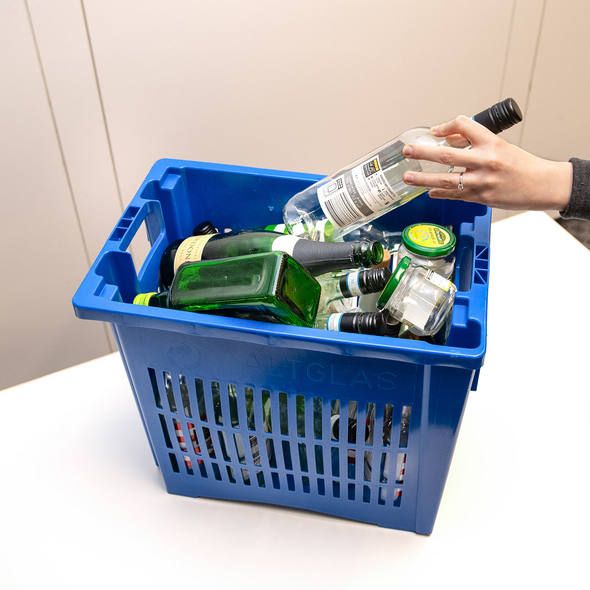 Ein blauer Kunststoffkorb enthält verschiedene leere Flaschen und Gläser. Eine Hand hält eine Flasche, die scheinbar zum Entsorgen in den Korb gelegt wird. Der Korb ist für die Sammlung von Glasflaschen gedacht.