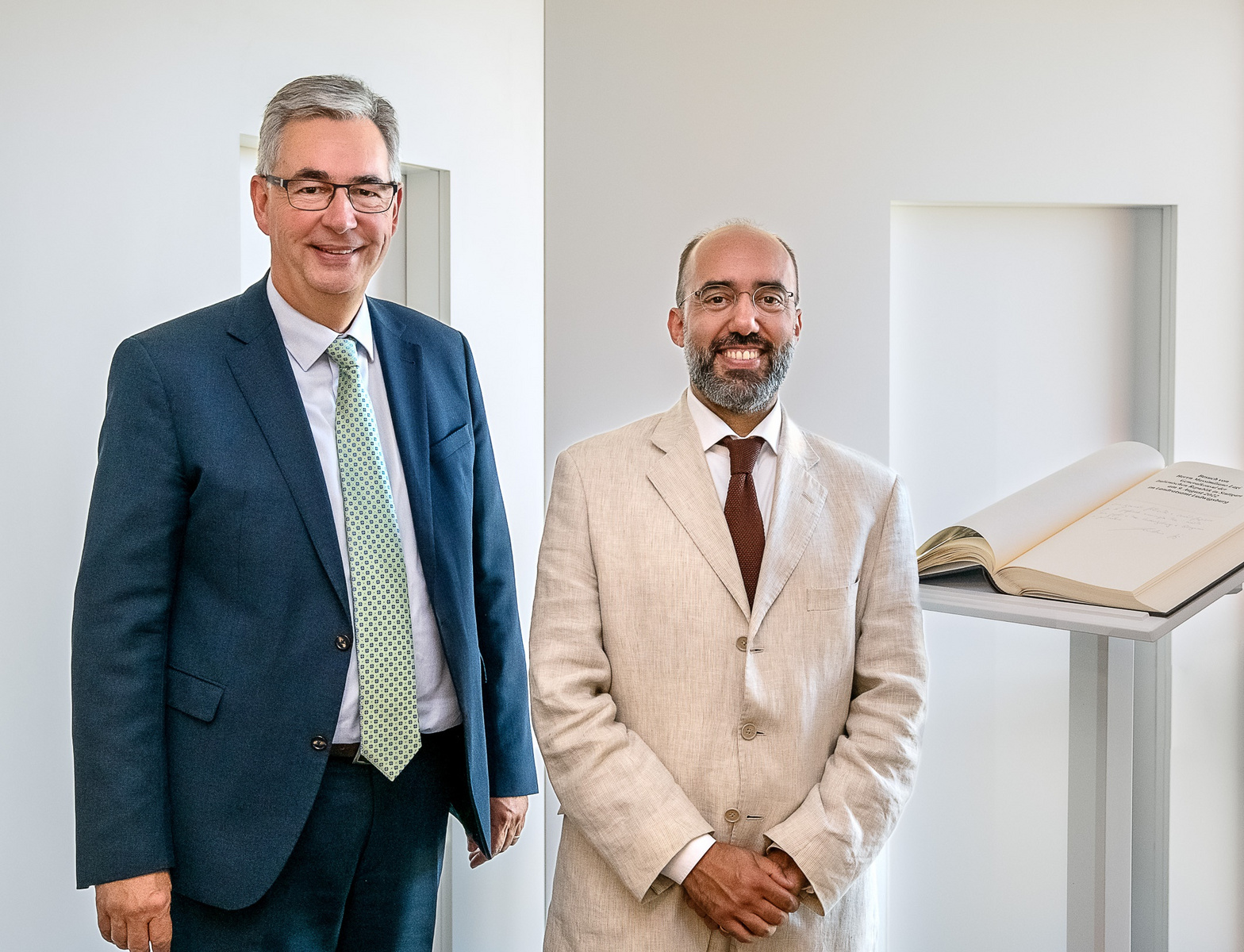 Zwei Männer stehen nebeneinander in einem hellen Raum. Der Mann links trägt einen Anzug und eine Krawatte, während der Mann rechts einen hellen Anzug ohne Krawatte anhat. Hinter ihnen ist ein offenes Buch auf einem Podest zu sehen.