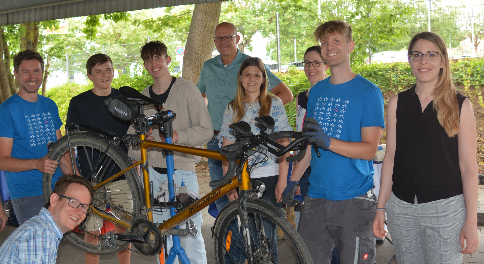 Eine Gruppe von acht Personen steht um ein Fahrrad, das auf einem Montageständer bearbeitet wird. Alle lächeln, einige halten Werkzeuge in der Hand. Die Szene scheint eine Gemeinschaftsaktivität zur Fahrradreparatur oder -wartung darzustellen.