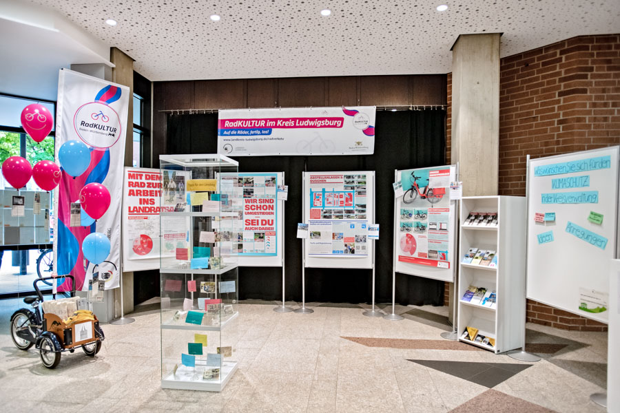 Eine Ausstellung zum Thema "Radkultur im Landkreis Ludwigsburg" mit Informationsständen, Flyern und einem Fahrradanhänger. Bunte Ballons sind verteilt, und es gibt zahlreiche Plakate, die zur radfreundlichen Mobilität aufrufen. Die Umgebung wirkt einladend und informativ.